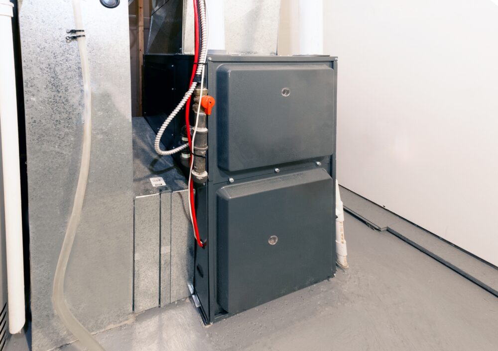 A gray residential HVAC furnace unit installed in a basement, surrounded by metal ductwork and electrical wiring, on a clean concrete floor.