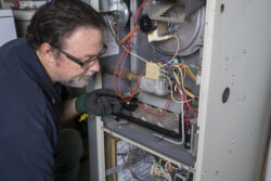 A technician wearing gloves inspects the wiring inside an open furnace with a flashlight.