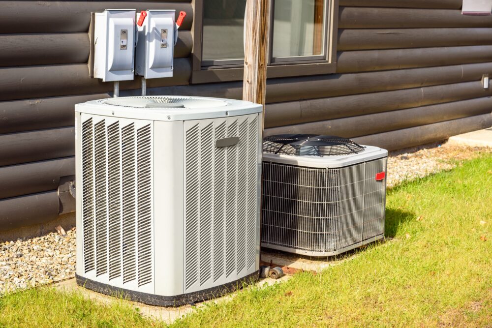 Two outdoor air conditioning units are positioned on concrete pads beside a building with wood siding.